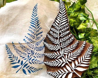 May include: A brown wooden block printing stamp featuring a carved fern leaf design, positioned next to a white fabric cloth displaying a blue ink-stamped fern leaf print. The stamp surface shows the raised white-painted fern pattern clearly.