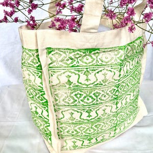 Può includere: Una borsa tote color crema con un design verde a fantasia. La borsa ha manici robusti ed è piena di fiori viola. Il motivo verde è un disegno ripetuto sulla parte anteriore della borsa.