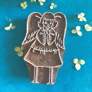 May include: A dark brown wooden printing block featuring a carved design of an angel with wings, a dress, and long thin legs. The carved lines are light in colour, creating a high contrast against the dark wood surface of the rectangular block.