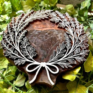 Könnte beinhalten: Ein runder Holzdruckblock mit einem tief geschnitzten, weißen Blumenkranzmotiv. Das Muster besteht aus zwei symmetrischen Blattzweigen, die sich unten treffen und mit einer einfachen Schleife zusammengebunden sind. Das Holz ist dunkelbraun.