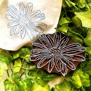 Könnte beinhalten: Ein dunkelbrauner Holzstempel mit einem geschnitzten Gänseblümchen-Motiv, platziert neben einem Stück cremefarbenem Stoff, der einen blauen Tintendruck desselben Gänseblümchens zeigt und das detaillierte Blumenmuster sowie die Textur hervorhebt.