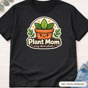 Op de afbeelding: Zwart T-shirt met een kleurrijke afbeelding van een lachende plant in een pot. Het ontwerp bevat de woorden "Plant Mom - crazy about plants", groene bladeren, een bruine pot en een witte achtergrond.