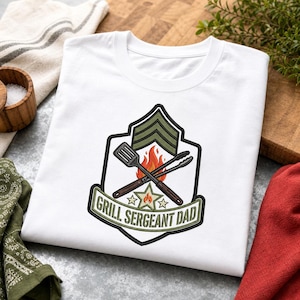 Op de afbeelding: Wit T-shirt met de tekst "Grill Sergeant Dad". De afbeelding toont een badge met sergeantstrepen, gekruiste tangen en spatel, vlammen en sterren. Een leuk cadeau voor de barbecue-liefhebber.