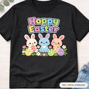 Op de afbeelding: Zwart T-shirt met de tekst "Hoppy Easter" boven drie cartoonkonijntjes. Elk konijntje heeft een andere kleur en houdt paaseieren en bloemen vast. Het ontwerp gebruikt pastelkleuren en is geschikt voor Pasen.