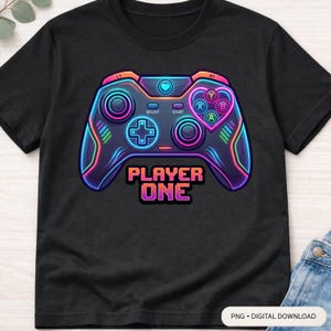 May include: Black t-shirt with a neon-coloured video game controller graphic. The controller design includes buttons, a directional pad, and the text "PLAYER ONE" in a pixelated font. The design has pink, blue, and orange accents.