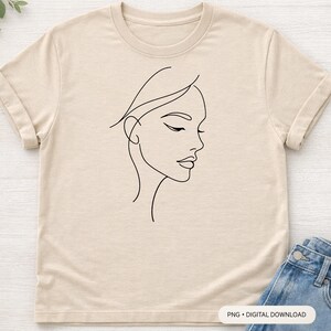 Op de afbeelding: Beige t-shirt met een minimalistische zwarte lijntekening van een vrouwengezicht in profiel. De afbeelding staat in het midden van de voorkant van het shirt. De mouwen zijn opgerold. Een blauwe spijkerbroek is rechtsonder zichtbaar.