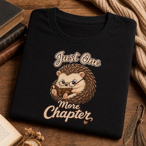 Op de afbeelding: Zwart T-shirt met een cartoon egel met een bril die een boek leest. De tekst "Just One More Chapter" staat boven en onder de egelillustratie. Het ontwerp is in bruin- en beigetinten.