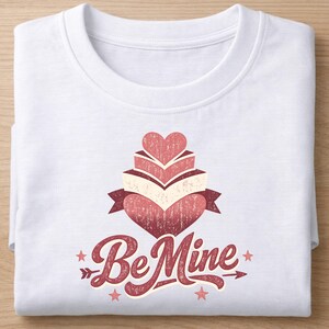 May include: White t-shirt with a red and cream-colored graphic. The graphic features a stack of hearts and books, a banner, stars, and the words "Be Mine" in a script font. The shirt is folded.