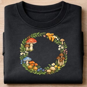 Op de afbeelding: Zwart sweatshirt met ronde hals en een aquarelkransontwerp van paddenstoelen, bladeren en bloemen. De krans bevat rode, oranje, blauwe en bruine paddenstoelen, met groene bladeren en witte en gele bloemen. Het sweatshirt is opgevouwen.