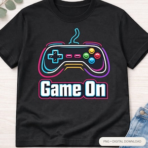 Könnte beinhalten: Schwarzes T-Shirt mit einem farbenfrohen Videospiel-Controller-Design und dem Text "Game On". Der Controller hat blaue, grüne, rote und gelbe Knöpfe. Ein lustiges Design für Gamer.