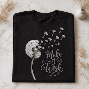 Op de afbeelding: Zwart T-shirt met een paardenbloemontwerp. De paardenbloem wordt afgebeeld met wegvliegende zaden en de woorden "Make A Wish" zijn in een sierlijk lettertype geschreven. Het T-shirt is netjes opgevouwen.