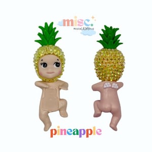 Puede incluir: Dos peque&ntilde;as figuras con forma de beb&eacute;s con cabezas de pi&ntilde;a. Las cabezas de pi&ntilde;a son amarillas y brillantes, con copas verdes y frondosas. Las figuras son de color canela claro y tienen un dise&ntilde;o caprichoso. La palabra "pineapple" est&aacute; escrita en letras coloridas.