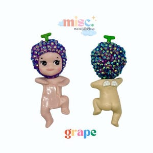 Puede incluir: Dos figuras decorativas, una rosa y otra beige, cada una con una cabeza en forma de uva adornada con gemas iridiscentes y un tallo verde. La palabra "grape" est&aacute; escrita en letras coloridas debajo.