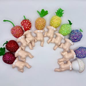 Puede incluir: Una colecci&oacute;n de adornos con forma de fruta enjoyados, que incluyen fresas, cerezas, pi&ntilde;as y un ar&aacute;ndano. Los adornos est&aacute;n dispuestos en semic&iacute;rculo con peque&ntilde;as figuras de color nude. La fruta est&aacute; cubierta de brillantes pedrer&iacute;a.