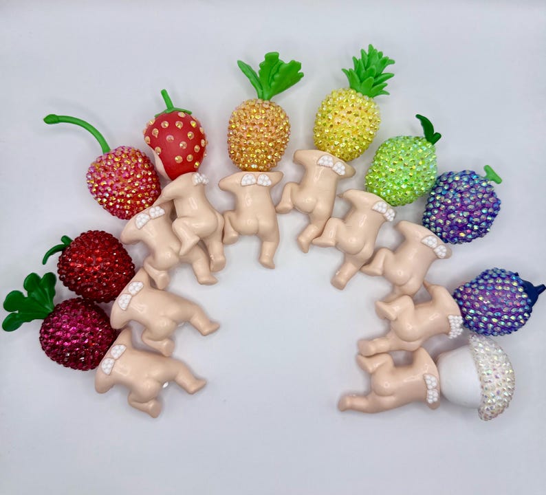 Puede incluir: Una colecci&oacute;n de adornos con forma de fruta enjoyados, que incluyen fresas, cerezas, pi&ntilde;as y un ar&aacute;ndano. Los adornos est&aacute;n dispuestos en semic&iacute;rculo con peque&ntilde;as figuras de color nude. La fruta est&aacute; cubierta de brillantes pedrer&iacute;a.