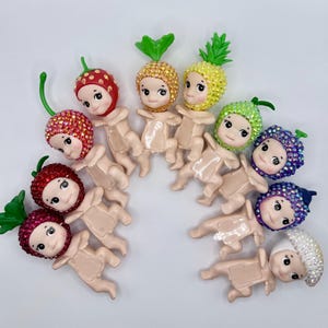 Puede incluir: Una colecci&oacute;n de figuras caprichosas, cada una adornada con un tocado con tem&aacute;tica de frutas. Las figuras presentan una variedad de frutas, incluyendo fresas, pi&ntilde;as y uvas, todas adornadas con acentos brillantes. Las figuras est&aacute;n dispuestas sobre una superficie blanca.