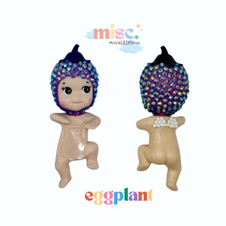 Puede incluir: Dos peque&ntilde;as y caprichosas figuras. Cada una tiene un cuerpo de tono de piel claro y una cabeza con forma de berenjena, cubierta de pedrer&iacute;a iridiscente. Una mira hacia adelante, la otra hacia atr&aacute;s. La palabra "eggplant" est&aacute; escrita en letras coloridas debajo.