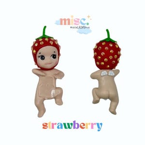Puede incluir: Dos peque&ntilde;as figuras, cada una con una cabeza en forma de fresa y un cuerpo desnudo. Las fresas son rojas con detalles dorados y tallos verdes. La palabra "strawberry" est&aacute; escrita en letras coloridas debajo.
