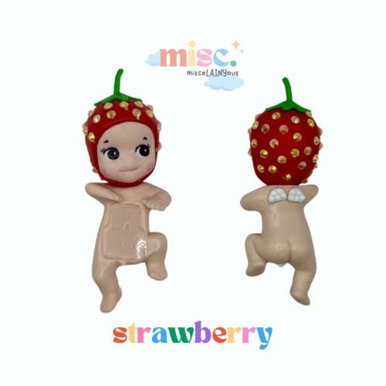 Puede incluir: Dos peque&ntilde;as figuras, cada una con una cabeza en forma de fresa y un cuerpo desnudo. Las fresas son rojas con detalles dorados y tallos verdes. La palabra "strawberry" est&aacute; escrita en letras coloridas debajo.