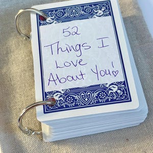 May include: A deck of playing cards held together by a silver ring. The top card has the handwritten text "52 Things I Love About You!" in purple ink. The cards have a blue decorative border.