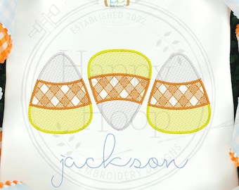 May include: White onesie with three embroidered candy corn designs and the name "Jackson" in blue cursive lettering. The onesie has a small tag with the text "Established 2022".