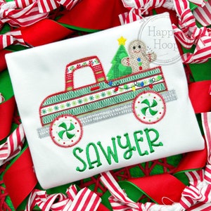 May include: White t-shirt with a Christmas truck design. The truck bed features a green Christmas tree, a gingerbread man, and string lights. The truck is decorated with red and green patterns. The name "SAWYER" is embroidered in green.