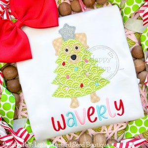 May include: A white onesie with a green and brown dog-shaped Christmas tree with lights and a silver star on top. The onesie is personalized with the name "Waverly" in red, pink, and blue letters.