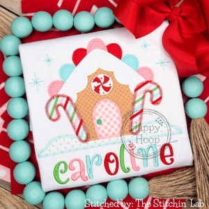 May include: A white onesie with a gingerbread house applique. The house is decorated with candy canes, a peppermint swirl, and a pink and white checkered door. The name "Caroline" is embroidered in colorful letters below the house.
