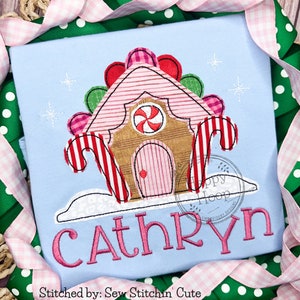May include: A light blue shirt with a pink and white candy cane house design and the name "Cathryn" embroidered in pink. The design includes candy canes, a gingerbread house, and a peppermint swirl.