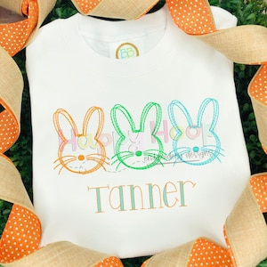 May include: White embroidered t-shirt featuring three colourful bunny outlines and the name "Tanner" in a light blue font.