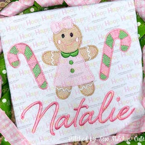 Puede incluir: Diseño bordado con una mujer de jengibre, un bastón de caramelo y el nombre "Natalie" en letras cursivas rosas. El diseño está sobre un fondo de tela blanca.