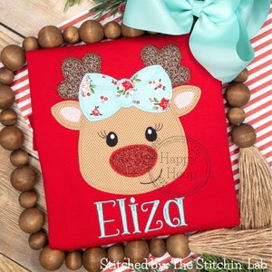 May include: Red Christmas shirt with a brown reindeer face with a blue bow and the name Eliza embroidered in blue.