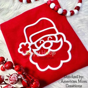 Puede incluir: Un artículo de tela roja con una cara de Papá Noel bordada en blanco. El diseño de Papá Noel incluye un gorro, barba y cara sonriente. También se ve el texto "Happy Hoop" y "Stitched by: American Mom Creations".