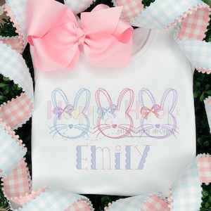 May include: White embroidered shirt with three bunny rabbits and the name "Emily" in pink and blue thread.