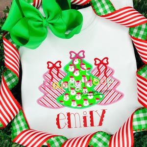 May include: A white onesie with a green Christmas tree design featuring Santa faces. The tree is surrounded by pink and red striped bows. The text "Happy Hoop Embroidery Designs" is printed below the tree. The name "Emily" is embroidered in white with a scalloped border below the design.