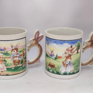 May include: Two white ceramic mugs with bunny rabbit handles. The mugs feature colorful illustrations of bunnies in a garden setting. One mug shows two bunnies near a fence, while the other shows a bunny holding a basket.