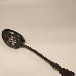 May include: A silver-toned metal serving spoon featuring an ornate, pierced lattice design within the bowl and intricate, embossed scrollwork detailing along the length of the handle, ending in a decorative, rounded terminal.