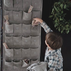 Può includere: Un calendario dell'avvento in lino grigio con 22 tasche numerate, ciascuna contenente un piccolo regalo. Un bambino sta raggiungendo una delle tasche per prendere un regalo.