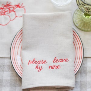 Puede incluir: Una servilleta de lino blanco con bordado rojo que dice "please leave by nine" está doblada y colocada en un plato rayado rojo y blanco.