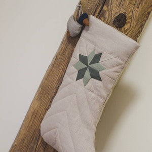 Peut inclure: Une chaussette de Noël matelassée beige avec un motif d'étoile verte et marron accrochée à une poutre en bois.