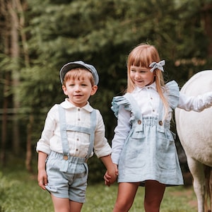Op de afbeelding: Twee kinderen, een meisje in een lichtblauwe jurk en een jongen in een wit shirt en lichtblauwe shorts, staan ​​naast een wit pony in een bosrijke omgeving. Het meisje aait de manen van de pony en de jongen kijkt naar de camera.