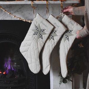 Könnte beinhalten: Drei weiße, gesteppte Weihnachtsstrümpfe mit grünen und weißen Sternmotiven hängen an einem Kaminsims. Die Strümpfe sind mit den Namen "Victoria", "Mark" und "Rose" personalisiert.