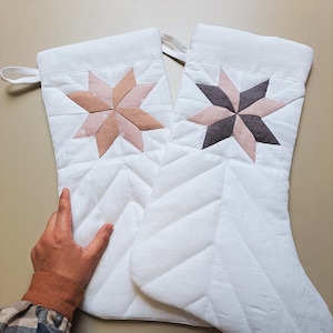 May include: Two white quilted Christmas stockings with a star design. One stocking has a pink and white star, and the other has a grey and white star.