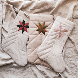 Könnte beinhalten: Drei beige gesteppte Weihnachtsstrümpfe mit Sternmustern in Rot, Grün und Rosa. Die Strümpfe haben ein diagonales Steppmuster und sind auf einem beigen Stoffhintergrund angeordnet. Jeder Strumpf hat eine Schlaufe zum Aufhängen.