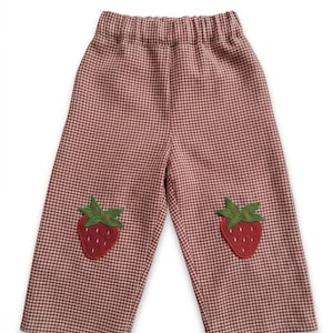 Può includere: Pantaloni con motivo a quadretti vichy marroni e bianchi e vita elastica. Ogni gamba presenta un'applicazione a forma di fragola rossa con foglie verdi centrata sul davanti. Il capo ha un taglio a gamba dritta e un orlo semplice.