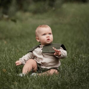 May include: A baby wearing a beige shirt, green shorts, and a dark green collar with a bird embroidery, sits in green grass. The baby has fair skin and light-colored hair, looking up with a curious expression.