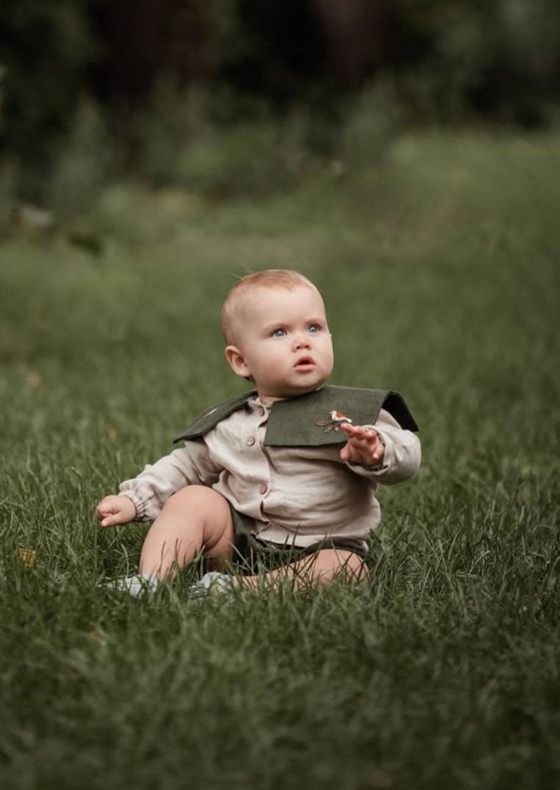 May include: A baby wearing a beige shirt, green shorts, and a dark green collar with a bird embroidery, sits in green grass. The baby has fair skin and light-colored hair, looking up with a curious expression.