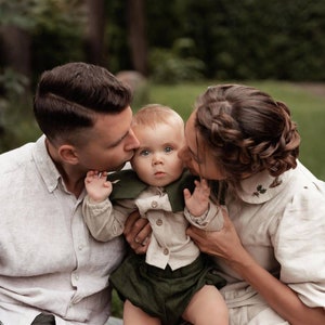 May include: A family portrait featuring a baby in a light-colored shirt and dark green shorts, with a matching green collar. The parents are kissing the baby's cheeks. The background is a blurred outdoor scene.