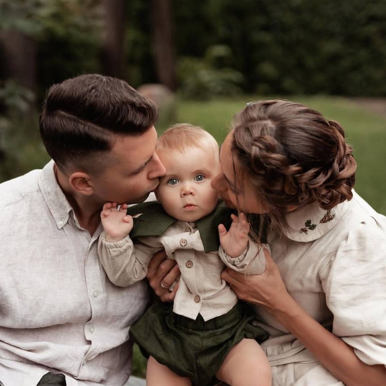 May include: A family portrait featuring a baby in a light-colored shirt and dark green shorts, with a matching green collar. The parents are kissing the baby's cheeks. The background is a blurred outdoor scene.