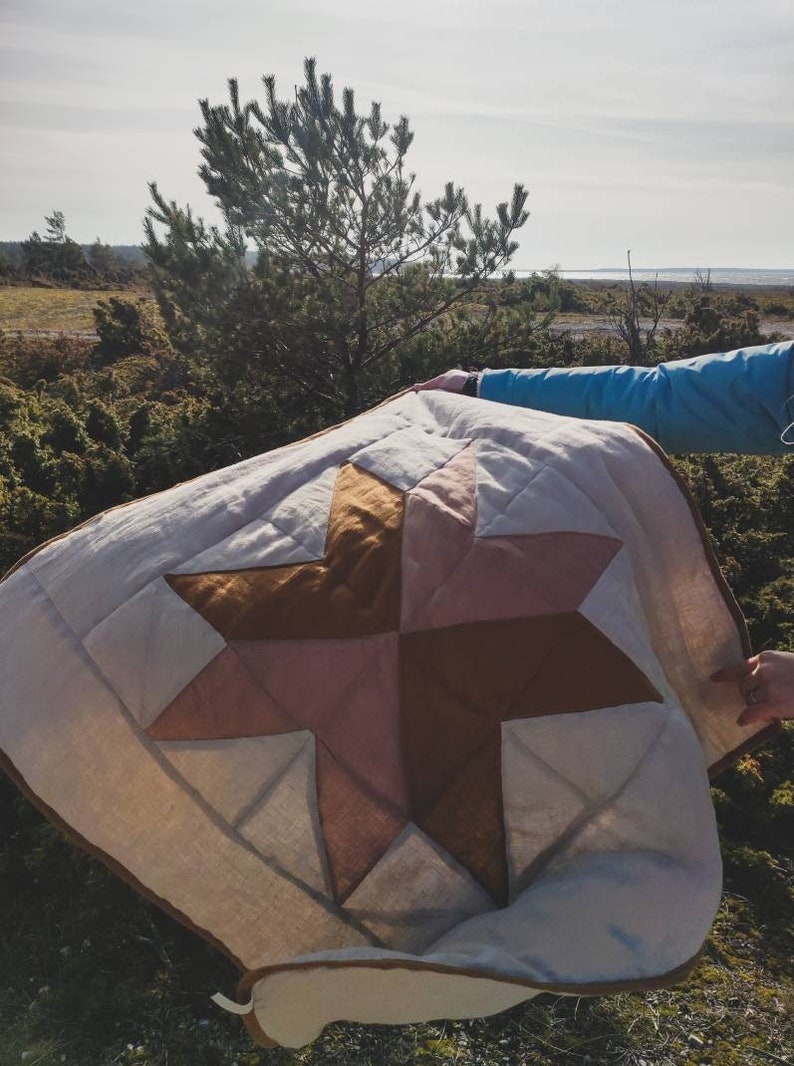 May include: A handmade quilt featuring a star design in shades of beige, pink, and brown. The quilt is held up outdoors, showcasing its geometric pattern and quilted texture. The quilt's edges are trimmed with a brown border.